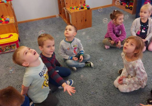 Dzieci siedząc na dywanie obserwują spadające bańki mydlane.